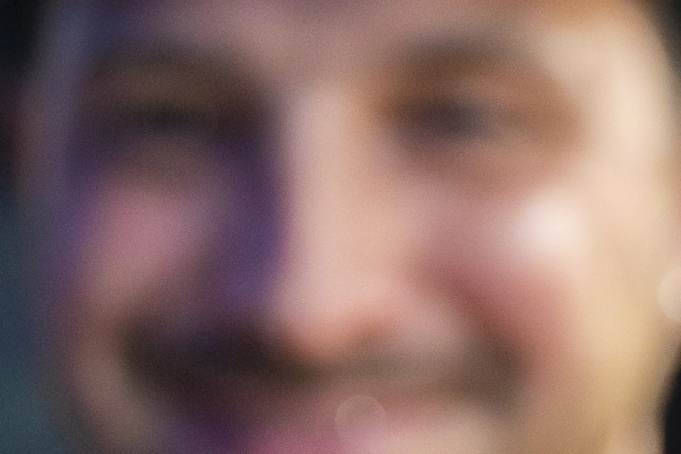 A person smiling holds a flaky pastry wrapped in a napkin, pictured in focus against a blurred background.