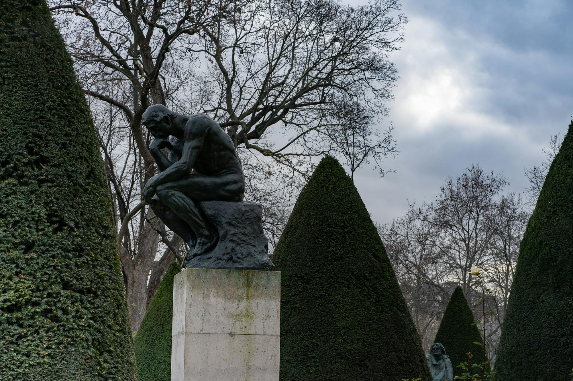다듬어진 원추형 수풀과 흐린 하늘 아래 잎이 없는 나무로 둘러싸인 받침대에 있는 로댕의 "사상가" 동상.