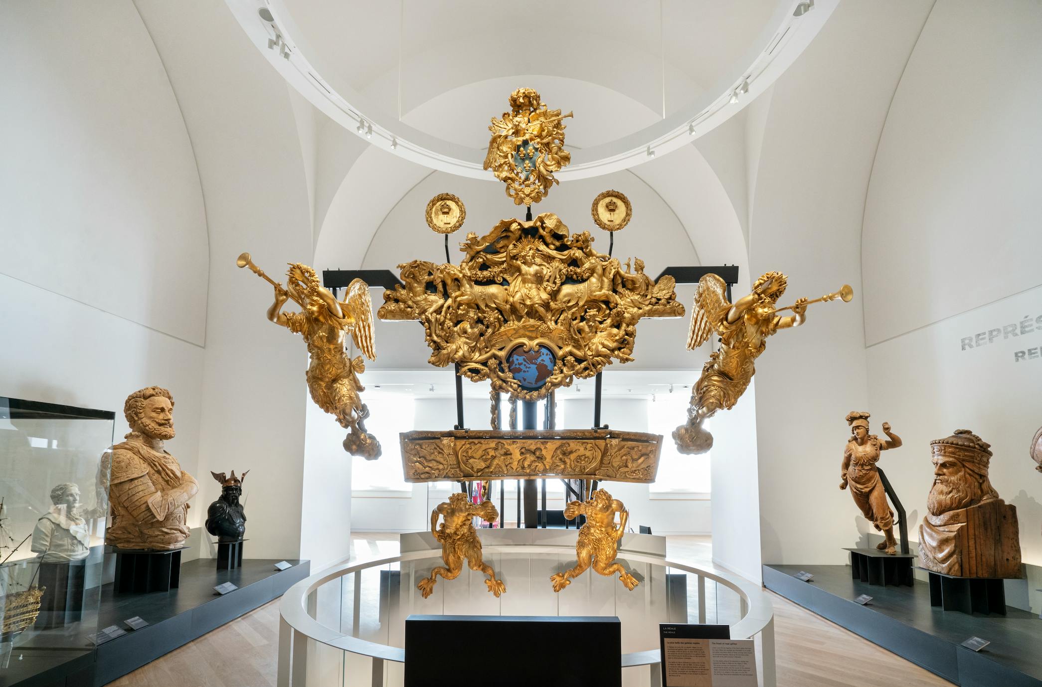 Ett galleri med en framstående gyllene skeppsnidning med keruber och trumpeterande änglar, omgiven av olika skulpturer.