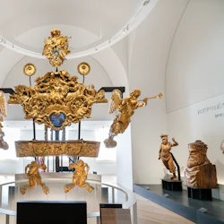 Ett galleri med en framstående gyllene skeppsnidning med keruber och trumpeterande änglar, omgiven av olika skulpturer.