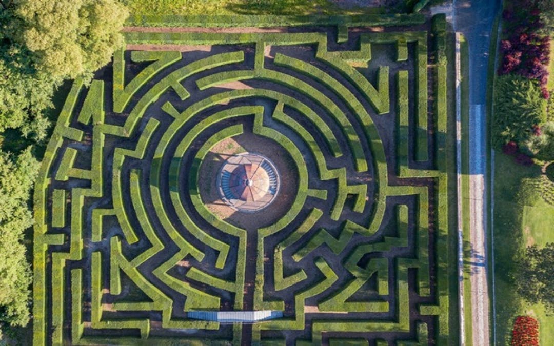 Aerial view of a circular hedge maze with pathways leading to a central paved area. Surrounding greenery is visible.