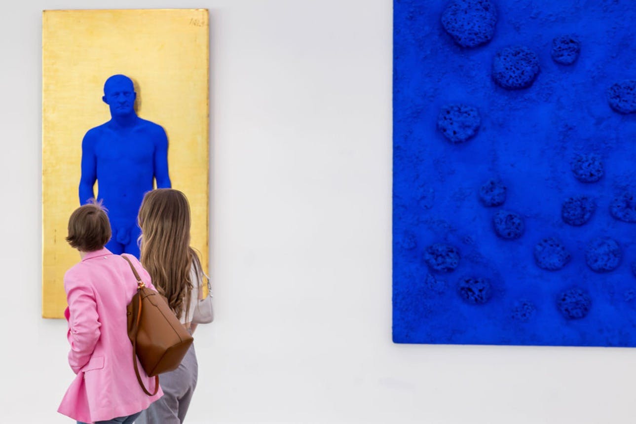 Dva lidé pozorují živé modré umělecké dílo lidské postavy na zlatém plátně v umělecké galerii.