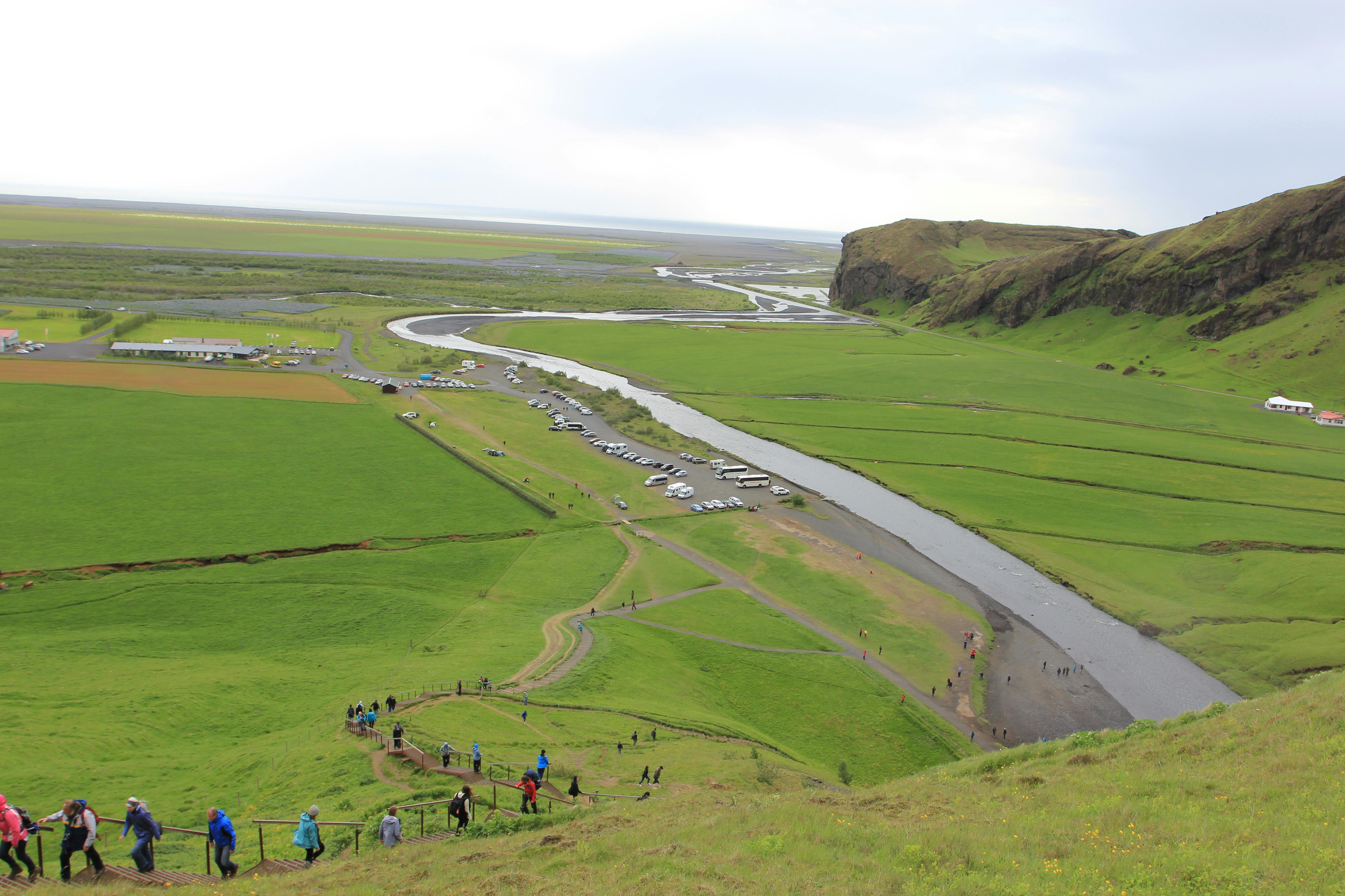 Aerial view of green fields with a winding river, parked cars, and people walking on paths leading up a hill.