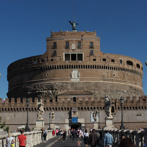 Large, circular brick fortress with a statue on top, viewed from a crowded pedestrian bridge adorned with statues under a clear blue sky.