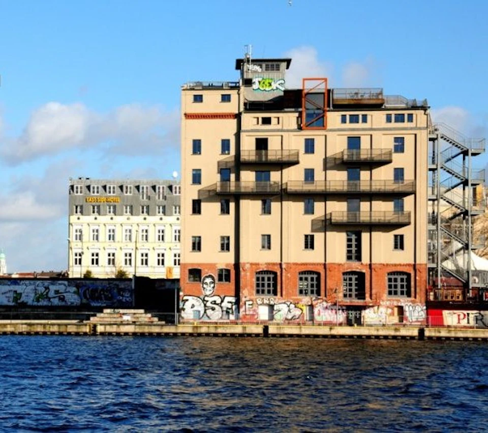 Three buildings by a waterfront with graffiti on the lower part of one. The sky is partly cloudy.