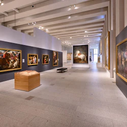 Konstgalleri med stora målningar i guldramar på grå väggar, en trämonteringslåda och modern belysning.