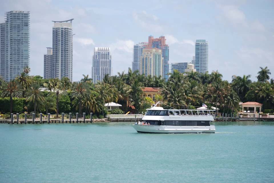A boat on turquoise water with high-rise buildings and palm trees in the background under a clear blue sky.
