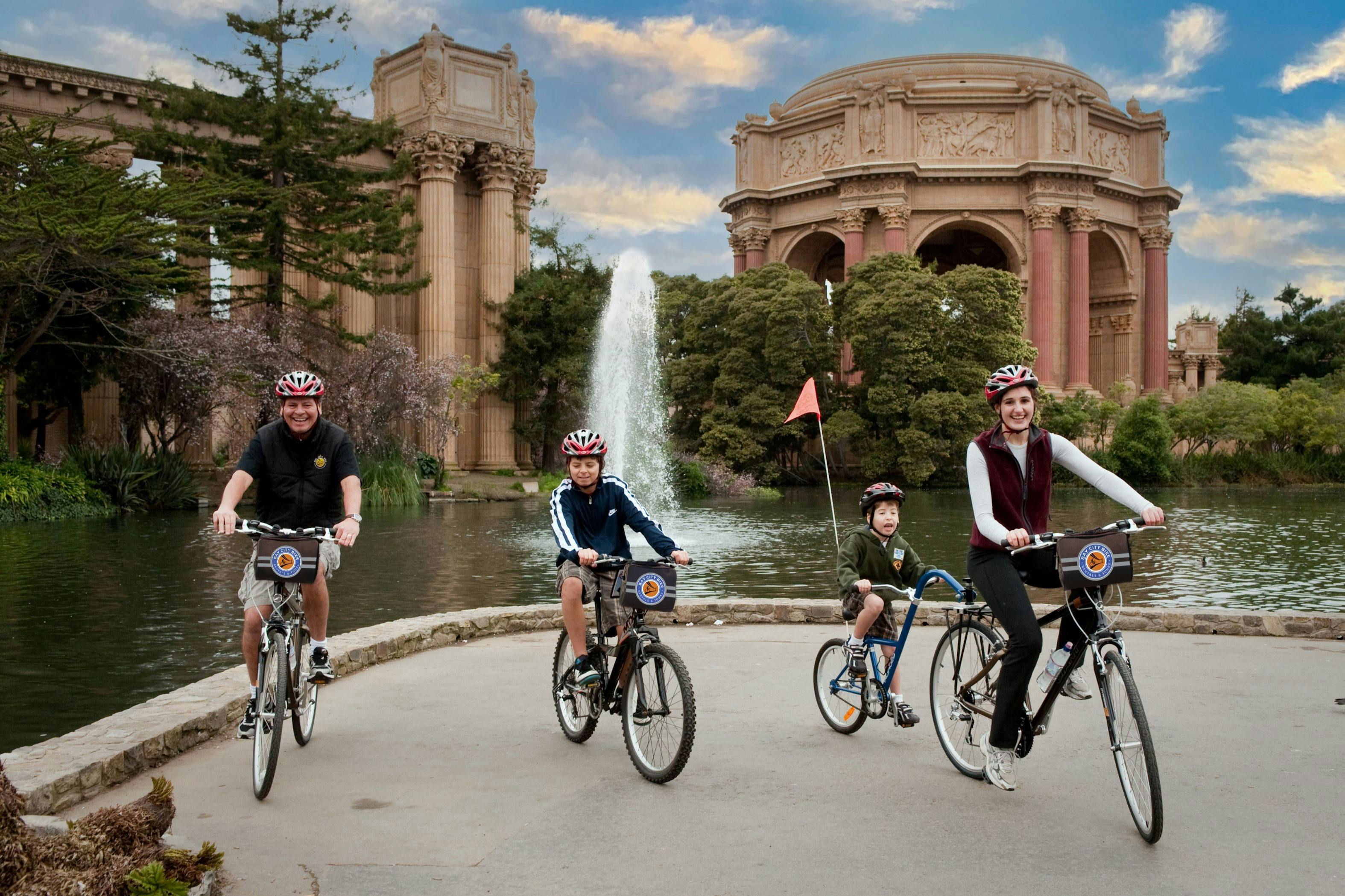 Three people on bicycles ride in front of a large building with columns, a fountain, and trees in the background.