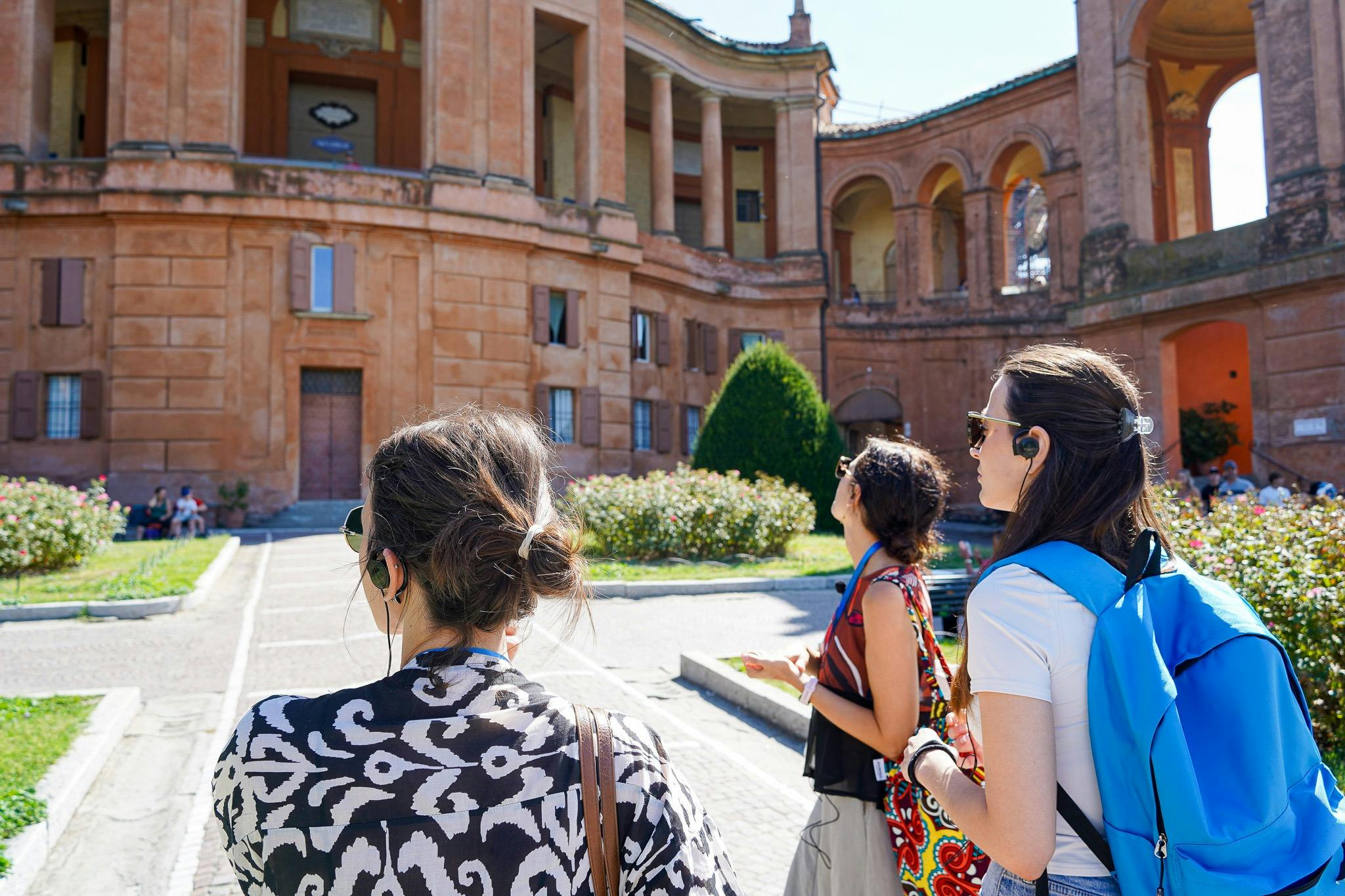 Three people with audio guides observe a historical building with columns and arches on a sunny day.