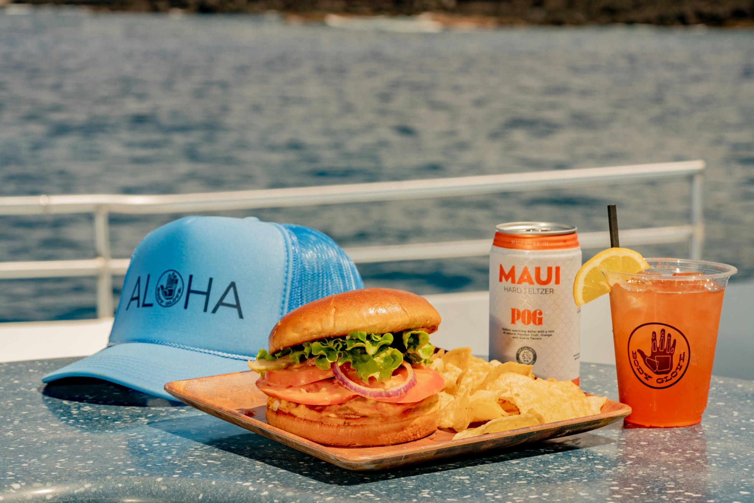 A sandwich with chips on a plate, a can of Maui Hard Seltzer, a drink with a lemon slice, and a blue hat on a table by water.