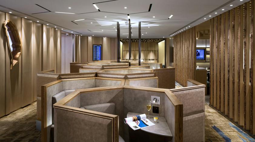 A modern lounge area with wooden partitioned seats, a table with a magazine and drink, and minimalistic lighting.
