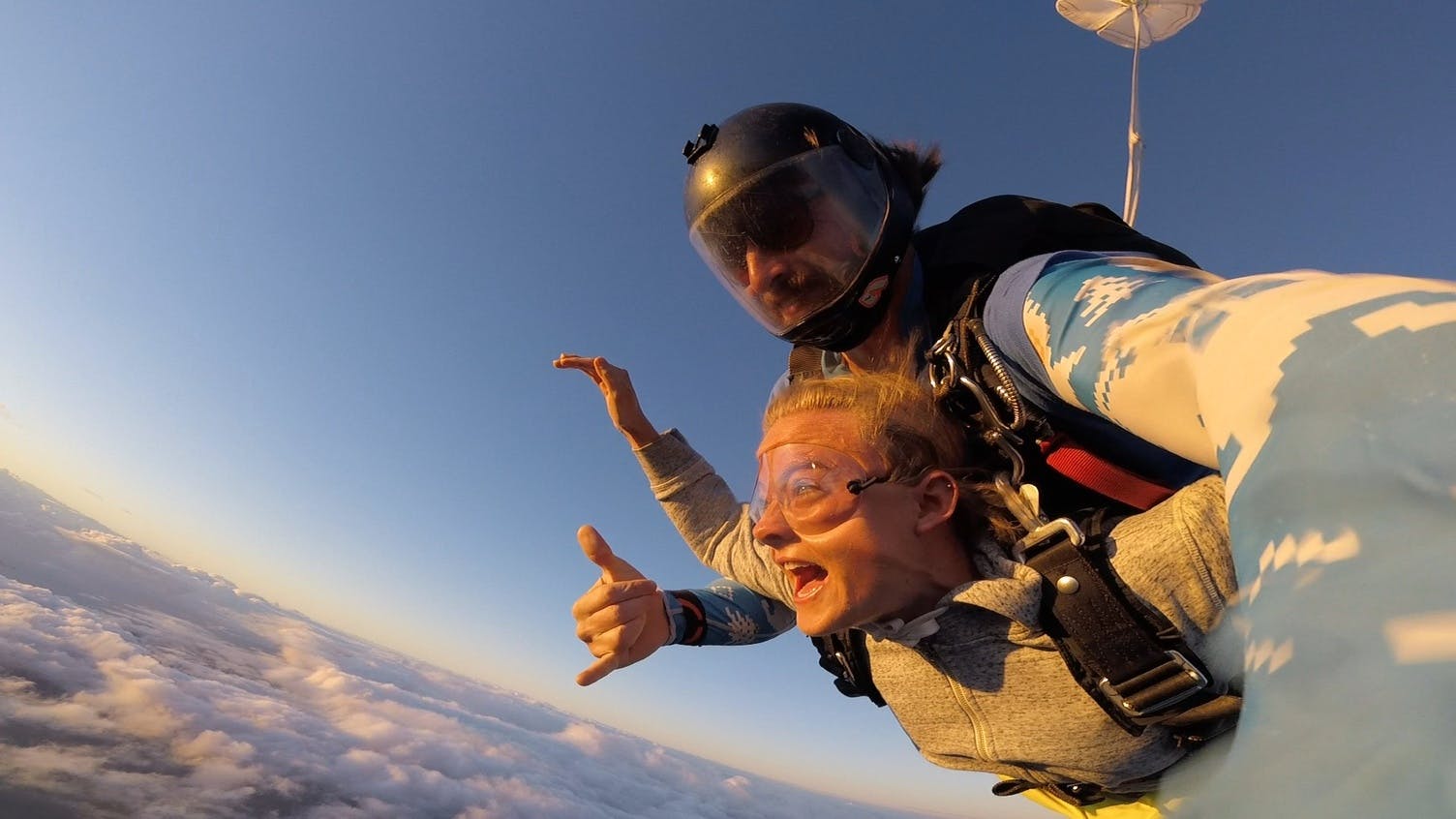 Due persone che fanno skydiving sopra le nuvole, indossando caschi e imbracature, con un paracadute aperto sullo sfondo.