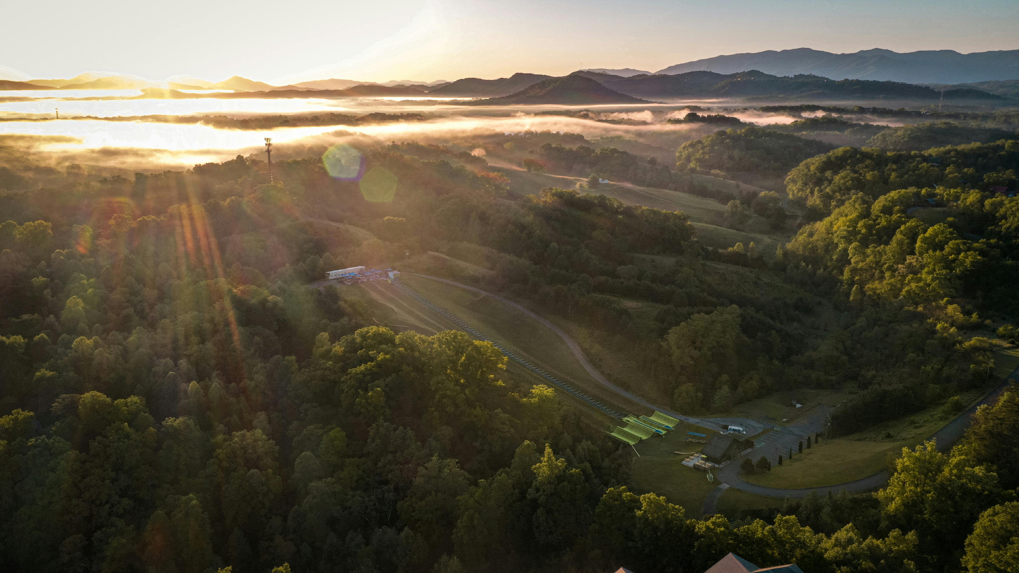 Veduta aerea di un paesaggio collinare illuminato dal sole con alberi, strade tortuose e una struttura con tetti verdi. La nebbia indugia sulle colline lontane.