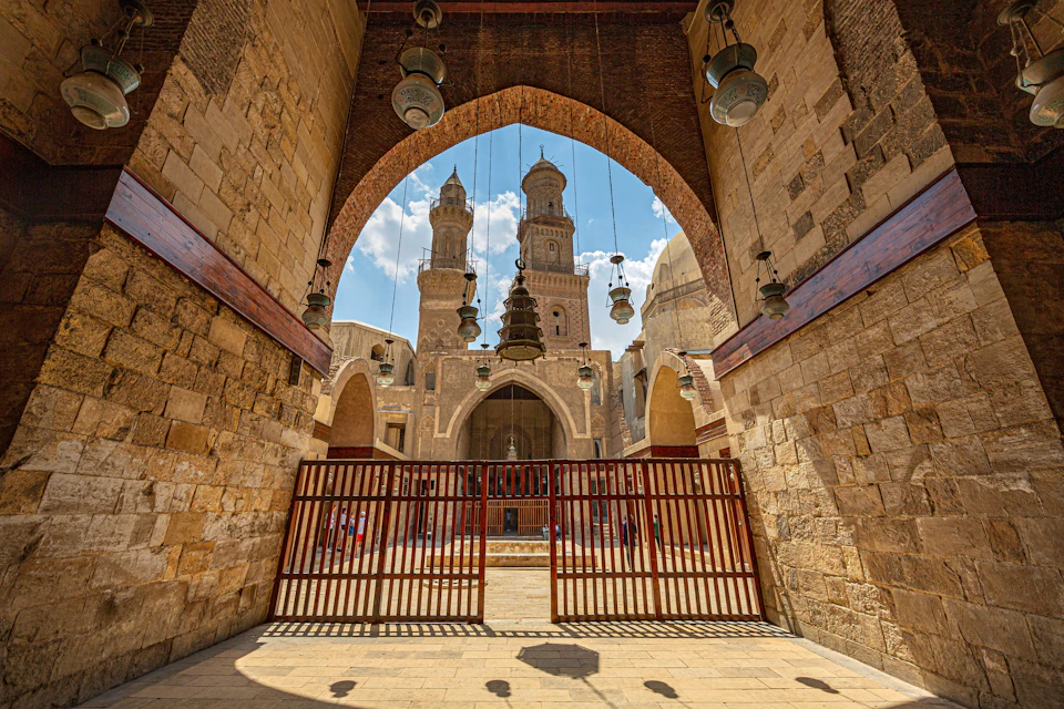 View of an architectural archway leading to an inner courtyard with two tall minarets and hanging lanterns.
