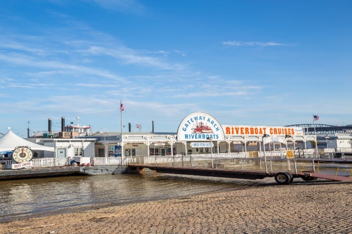 Αποβάθρα ποταμόπλοιων με πινακίδες "Gateway Arch Riverboats" και "Riverboat Cruises", κοντά σε ξύλινο διάδρομο και αμμώδη παραποτάμια περιοχή.