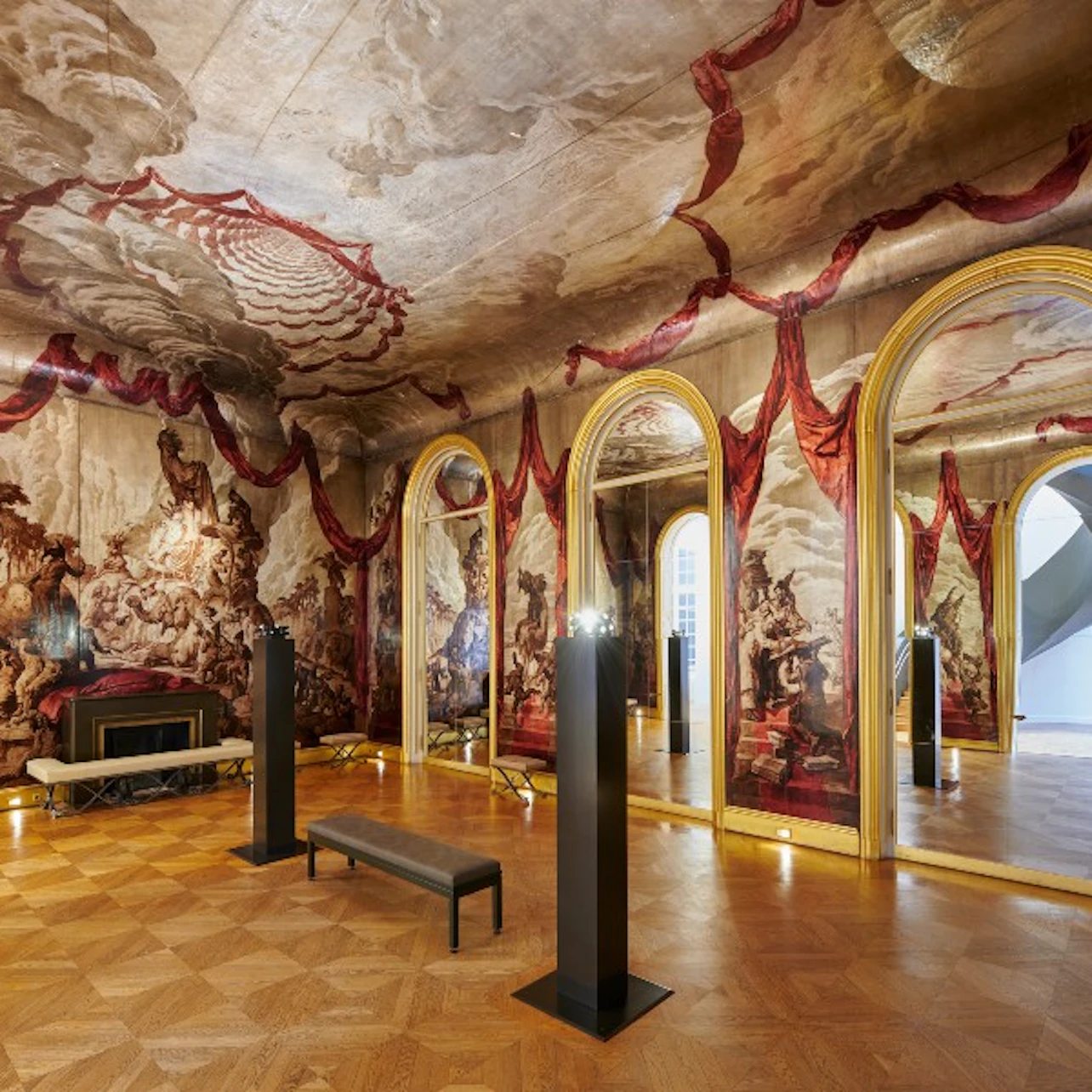 Ornate room with large ceiling and wall murals, gold-framed mirrors, a bench, and columns. Parquet flooring and historical decor.