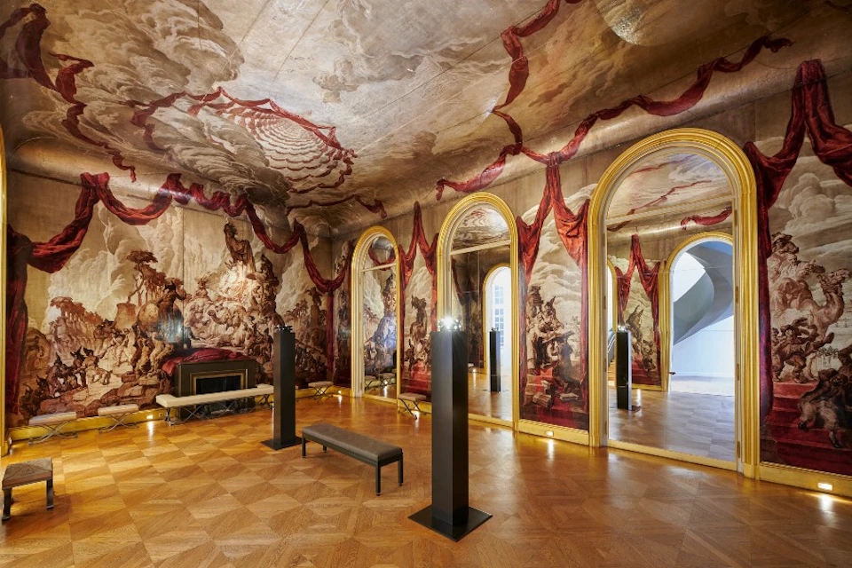 Ornate room with large ceiling and wall murals, gold-framed mirrors, a bench, and columns. Parquet flooring and historical decor.