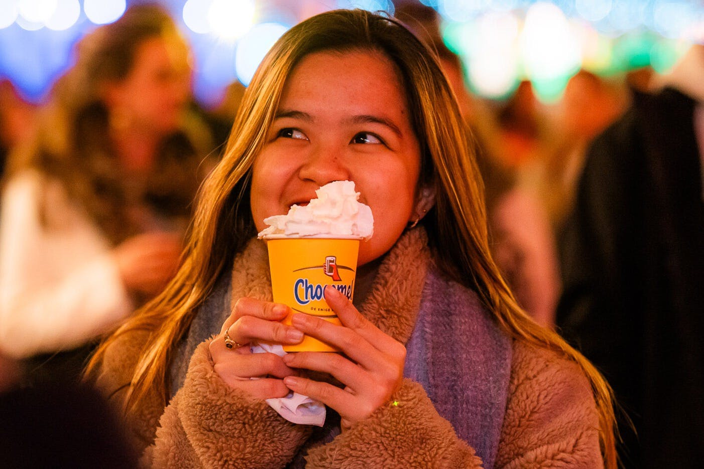 A woman in a brown coat smiles while holding a yellow cup filled with whipped cream, surrounded by bright, colourful lights.