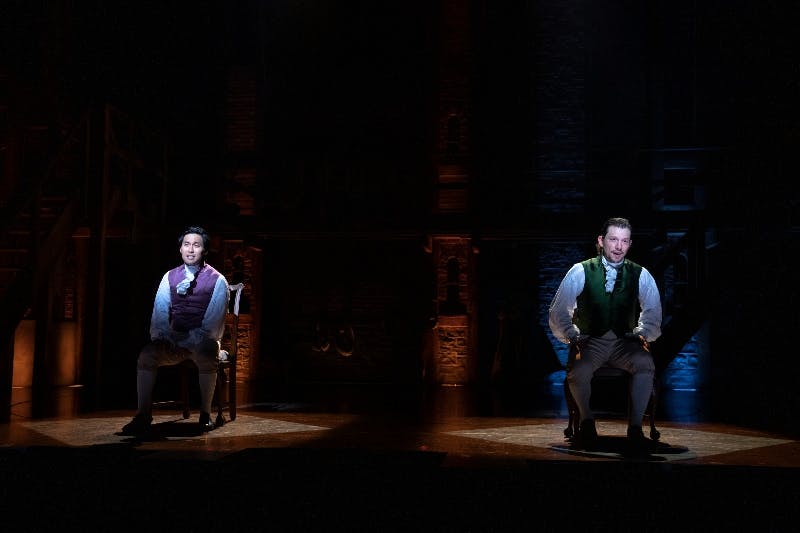 A person in period costume sits on a chair under a spotlight on a dimly lit stage. Brick wall and stairs are in the background.