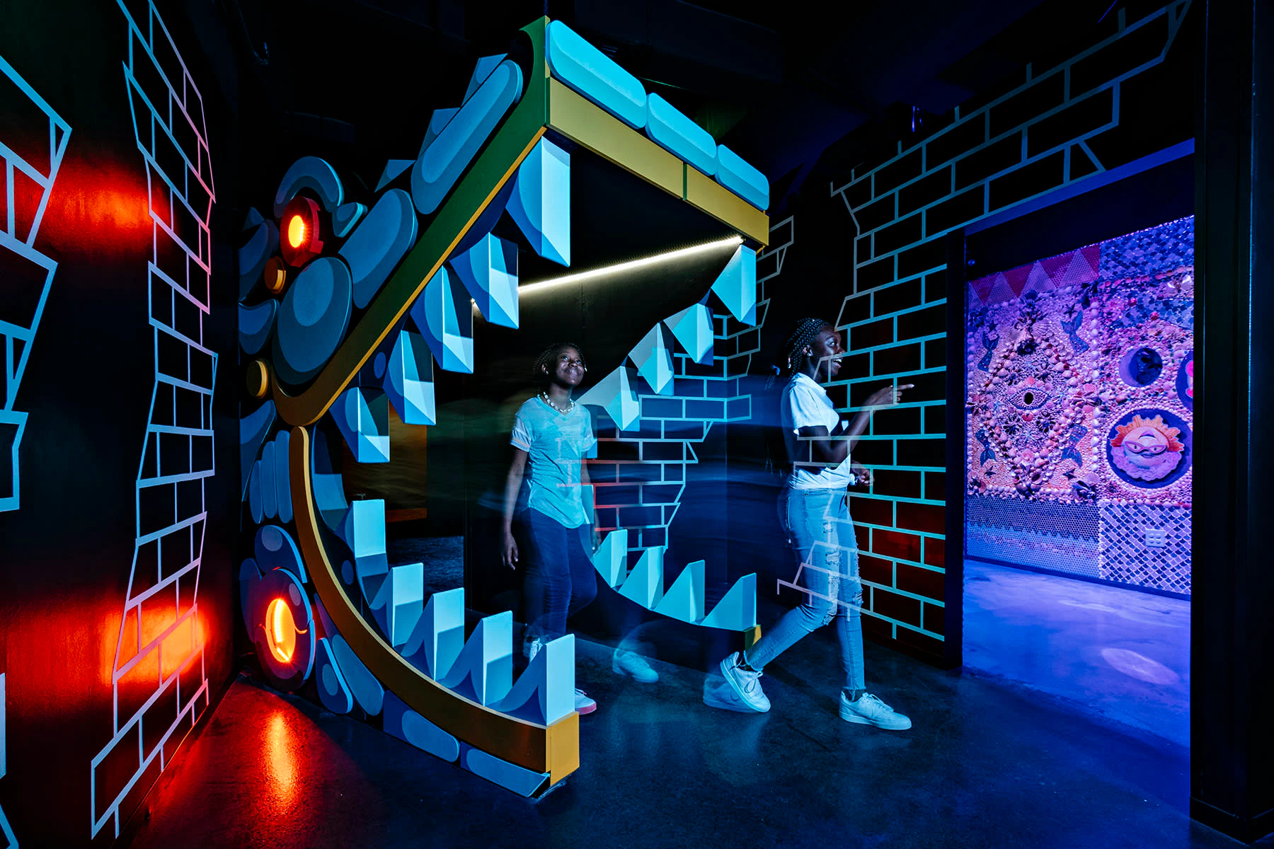 Two people walk through a large, cartoonish open-mouthed dragon display in a dimly lit, colorful exhibit space.