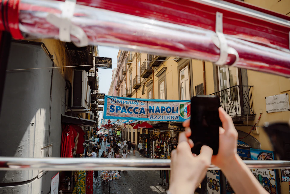 A person holds a smartphone to take a photo of a bustling street in Naples, Italy, with a banner overhead reading 'Benvenuti a Spacca Napoli.'