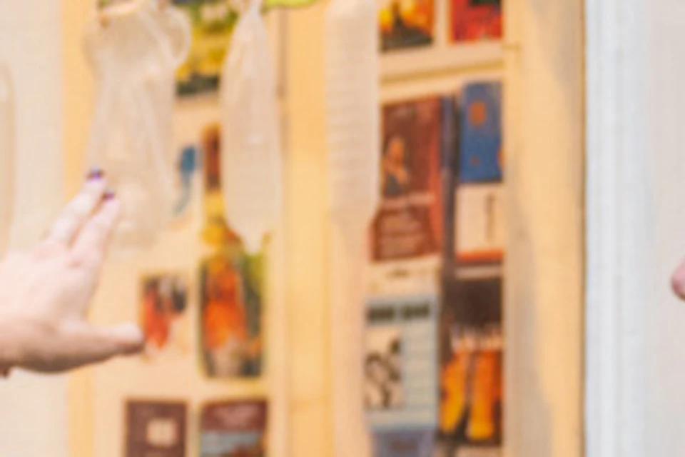 Two people look at a shop display of colorful condoms and various small items. Another person gestures towards the display.