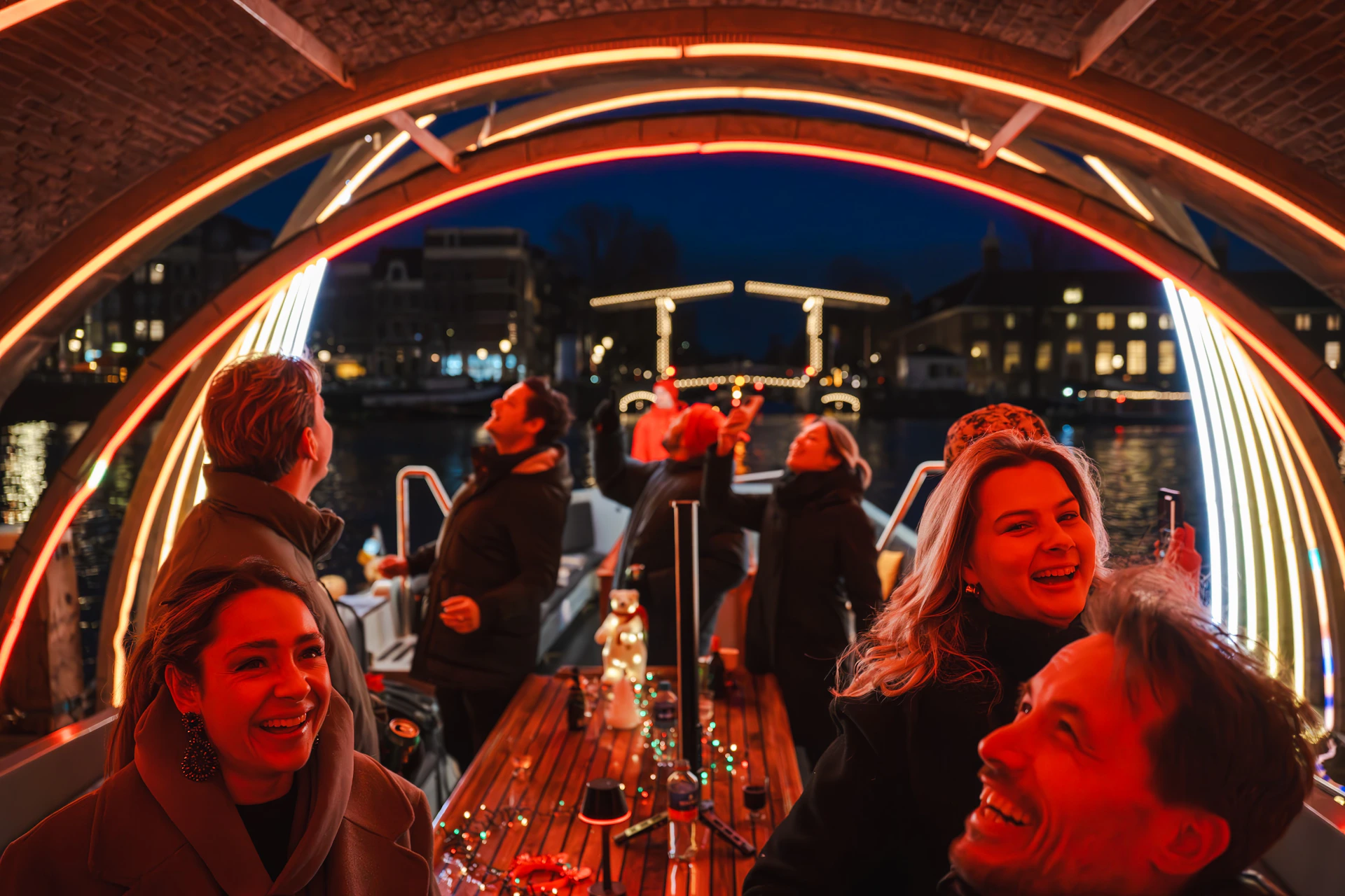 A group of people enjoying an evening on a lit-up boat under an arch, with a lighted bridge in the background.