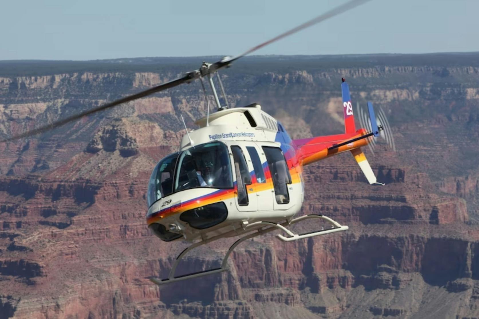 A helicopter with colorful markings flying over a canyon landscape under a clear sky.