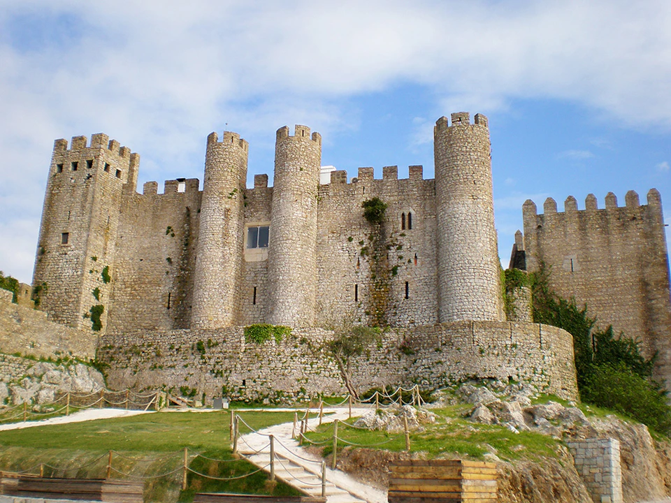 Stone castle with circular towers, crenelated walls, and small windows, surrounded by a grassy area and ropes. Blue sky overhead.