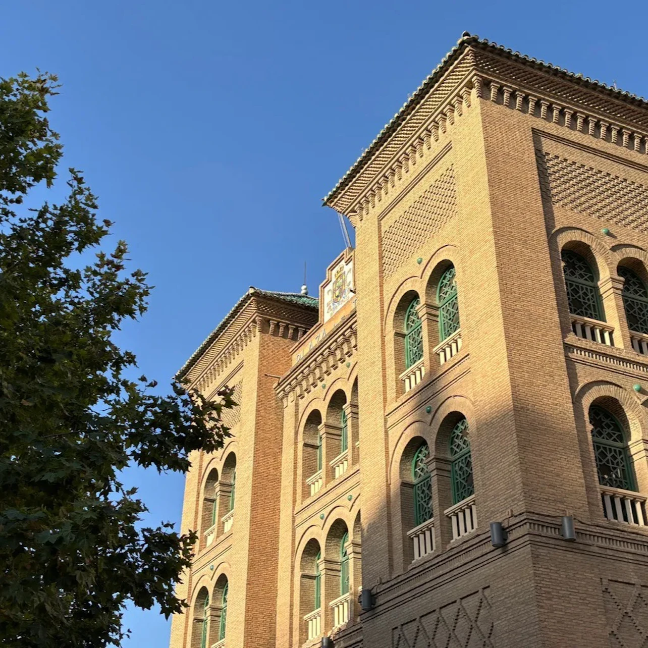 Brick building with ornate windows under a clear blue sky, partially obscured by a tree.