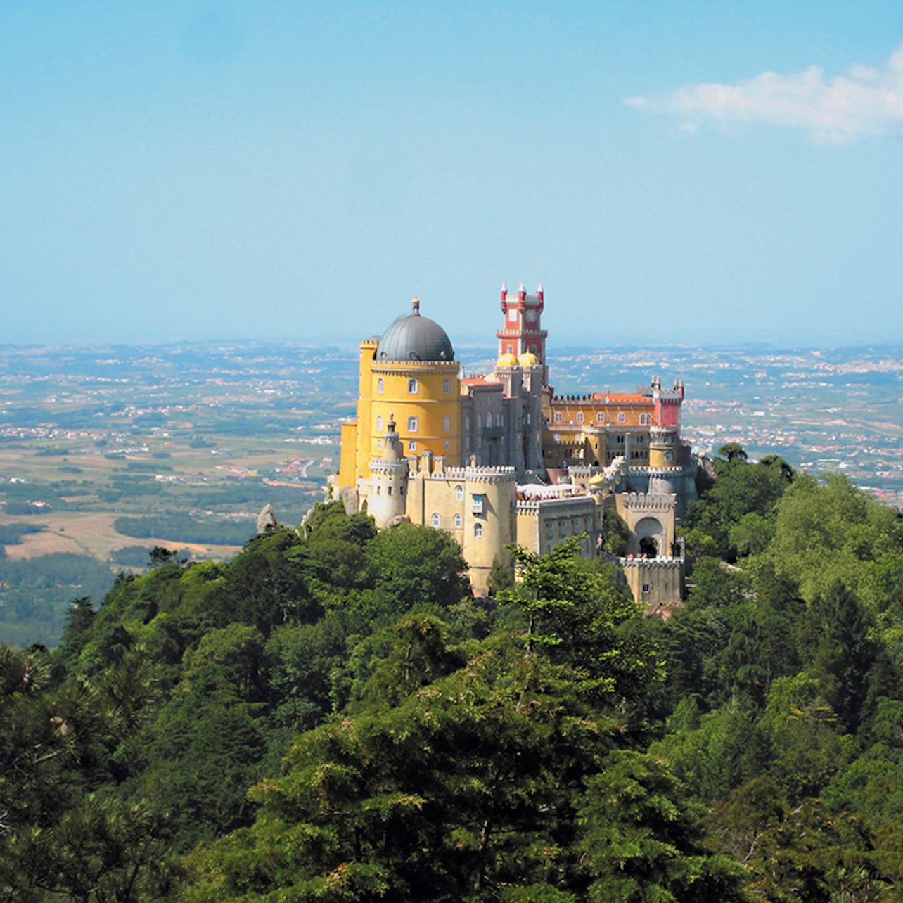 A colorful, historic castle situated atop a forested hill, overlooking a vast, sprawling landscape under a clear blue sky.