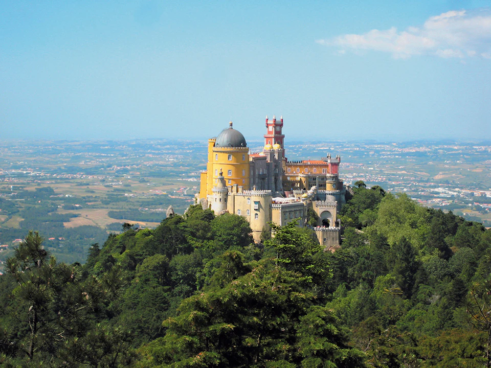 A colorful, historic castle situated atop a forested hill, overlooking a vast, sprawling landscape under a clear blue sky.