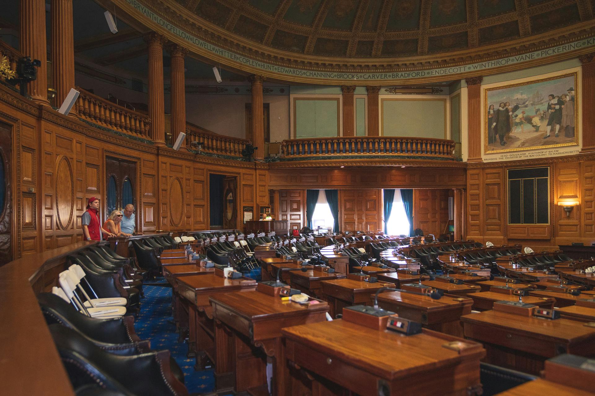 A legislative chamber with empty desks and seating, detailed woodwork, and a domed ceiling. Two people stand at the back.