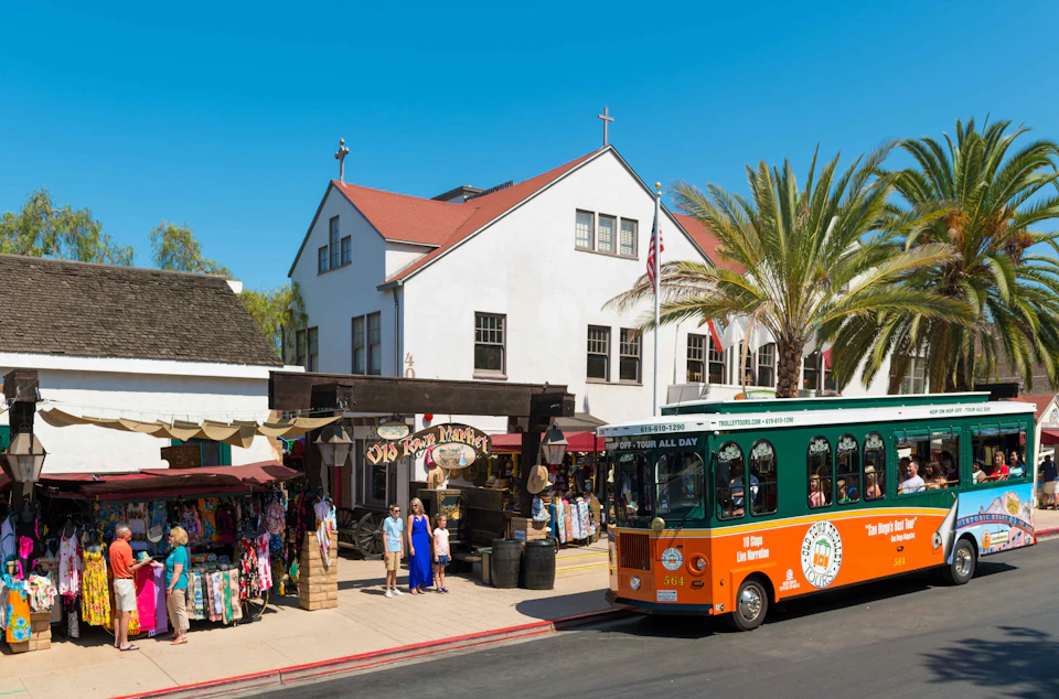 A green trolley bus passes by a white building and market shop selling colorful clothes. People stand and browse outside.