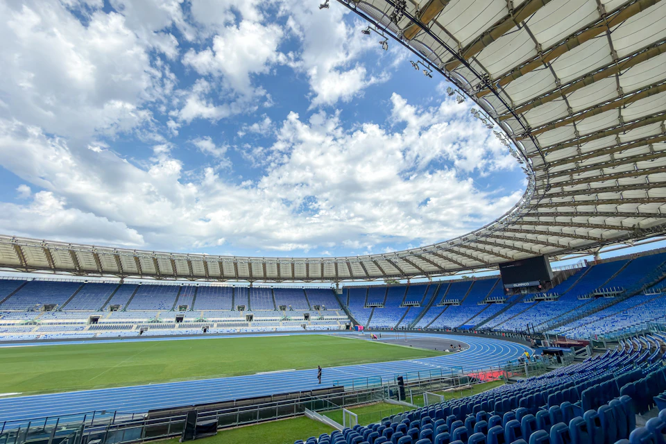 A large outdoor stadium with a green field, blue running track, tiered seating, and a partly cloudy sky above.