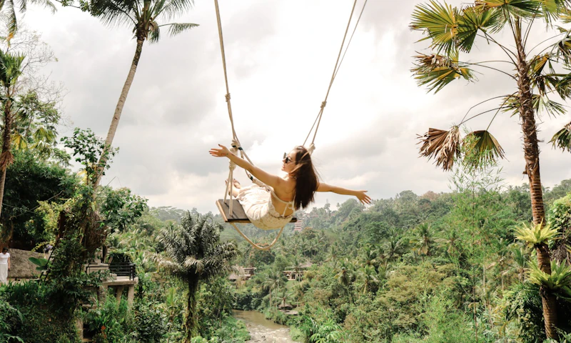 A woman on a swing over a lush green valley with palm trees, wearing a white dress and sunglasses, arms outstretched.