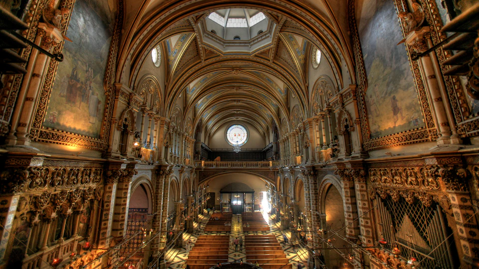 Interior of an ornate church with arched ceilings, a large circular stained-glass window, and rows of wooden pews in symmetry.