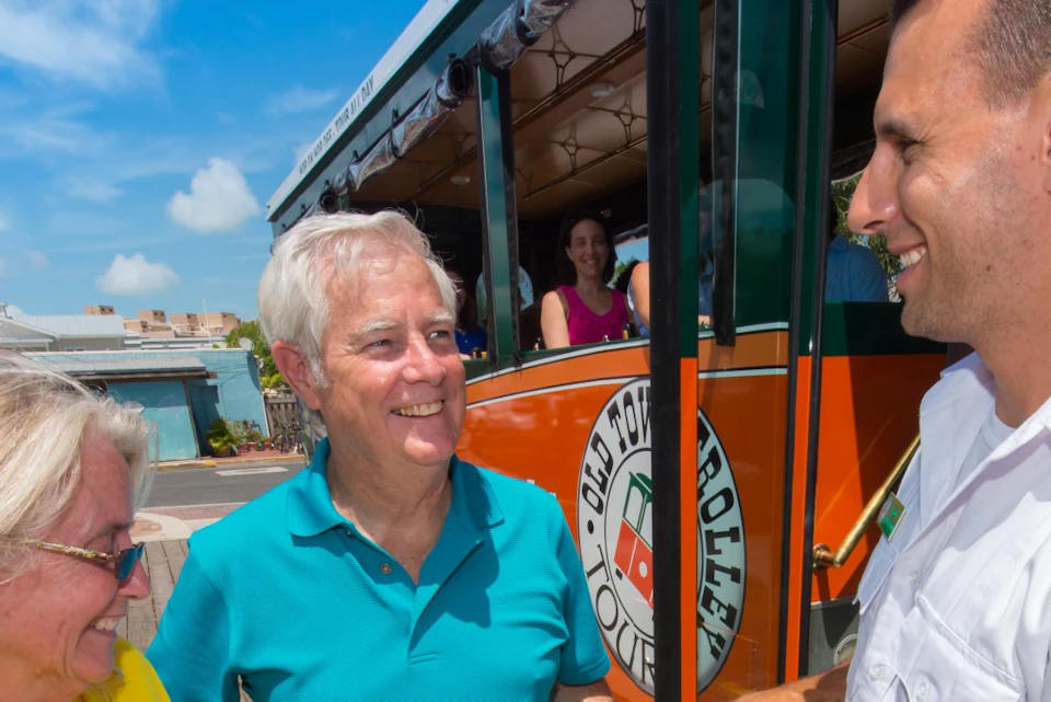 A couple stands outside an orange trolley, receiving tickets from a smiling employee in uniform under a sunny, blue sky.