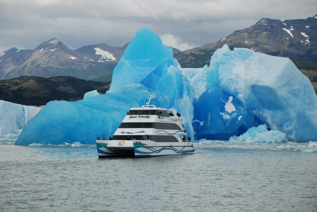A tour boat on a lake near large blue icebergs with mountains in the background under a cloudy sky.