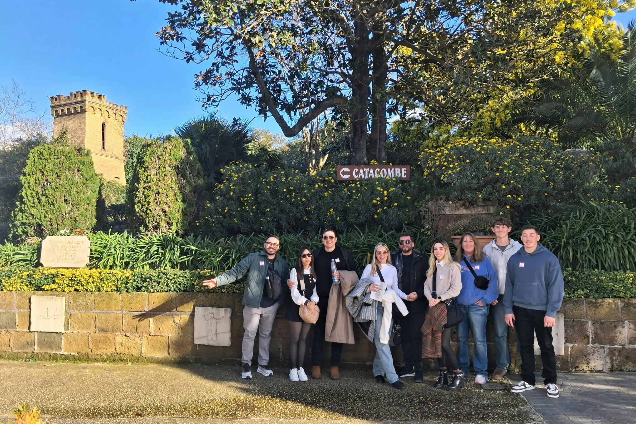 Un groupe de neuf personnes posant en plein air devant un panneau indiquant « Catacombe », au milieu des arbres et de la verdure.