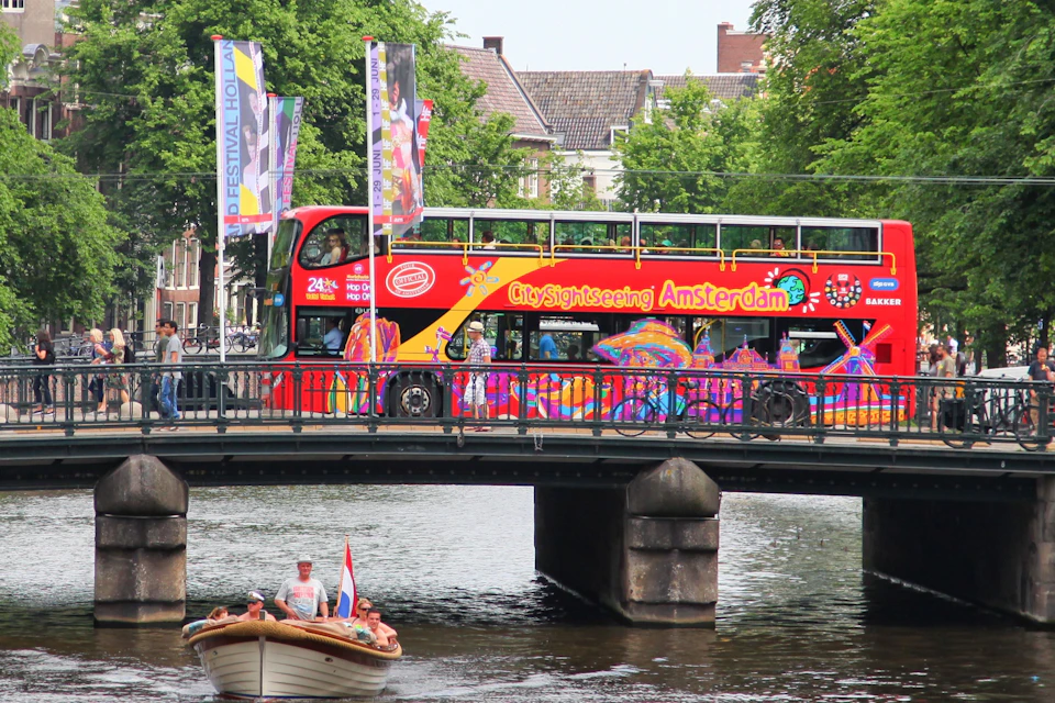 A red sightseeing bus with colorful designs crosses a bridge over a canal in Amsterdam, with trees and buildings in the background.