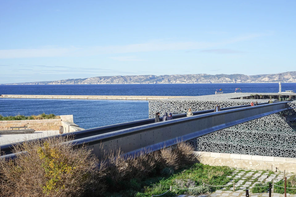 People walking on a bridge with intricate lattice sides, overlooking the sea and distant mountains under a clear blue sky.