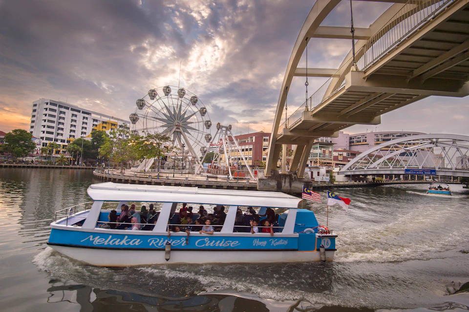 A boat with "Melaka River Cruise" written on it sails under a bridge; a ferris wheel and buildings are in the background.
