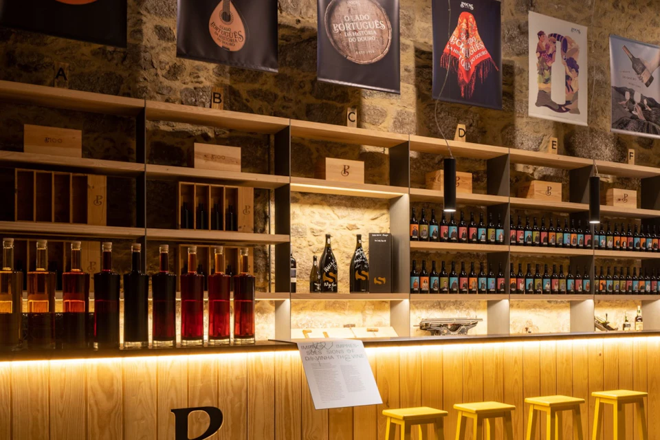 A wine shop interior with shelves of bottles, wooden bar counter, yellow stools, and decorative posters on the wall.