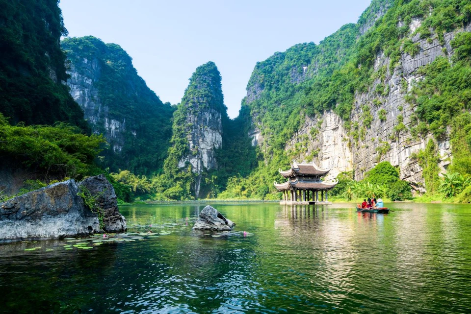 Serene lake with a traditional pavilion on stilts, surrounded by tall, lush green mountains under a clear blue sky.