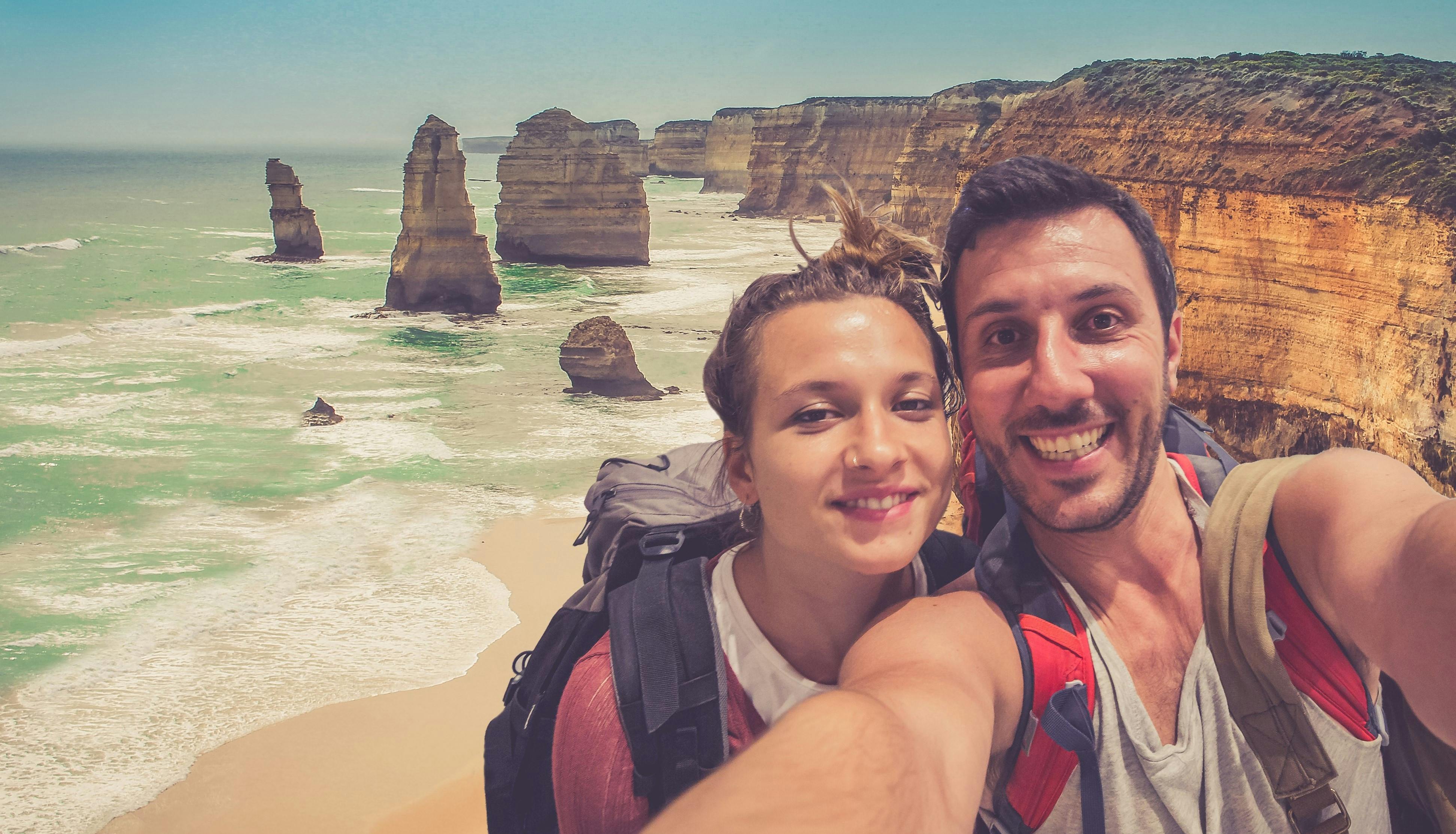 微笑むカップルがバックパックを背負った自撮り写真を撮り、背の高い階層の崖が特徴的な険しい海岸線の前に立っています。