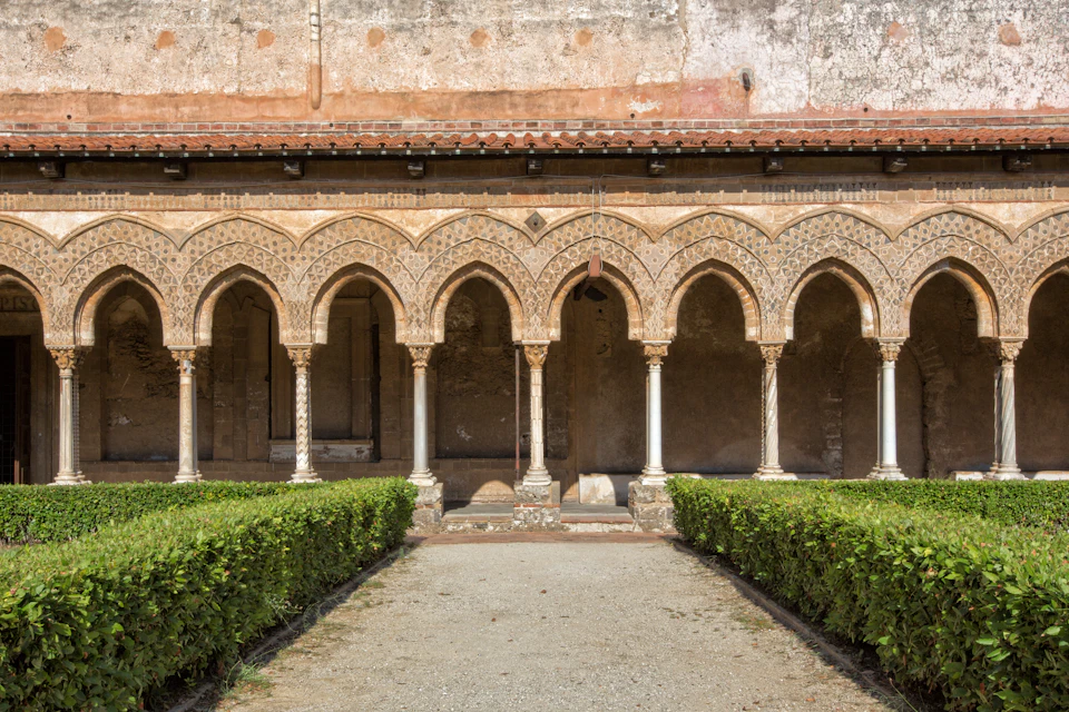 Row of ornate stone arches and columns with a tiled roof above and manicured green hedges below, in a historical courtyard setting.