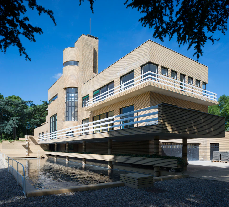 Modern, multi-story building with tan brick exterior, large windows, and balconies. Reflective pool in the foreground.