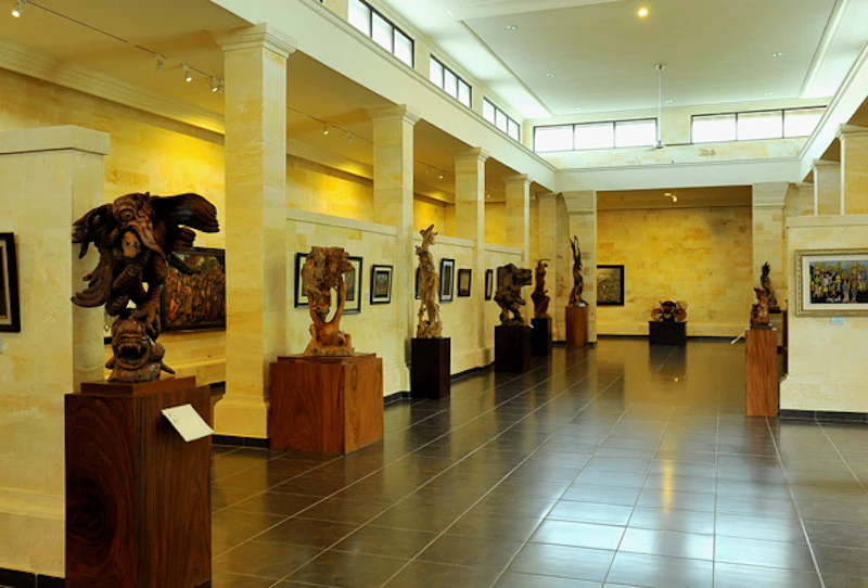 An art gallery featuring sculptures on pedestals and framed artworks on walls, with tiled flooring and natural lighting.