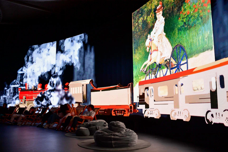 People sit in a dark room with large screens displaying a moving train and a woman on a bicycle in an artistic style.